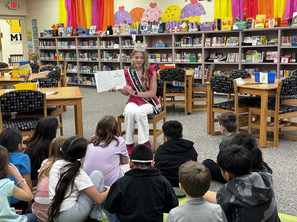 A guest reader seated in front of third graders reading a book she wrote when she was 14 in the media center.
