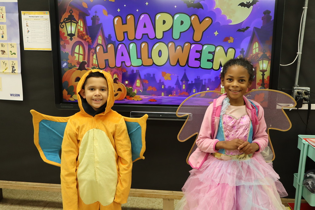 Two students dressed up in costumes standing in front of a Happy Halloween sign