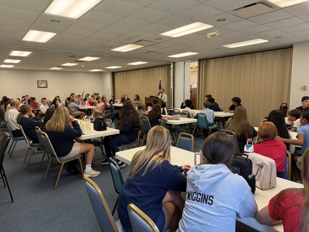 students seated at tables listening to a woman speaking to them from a podium