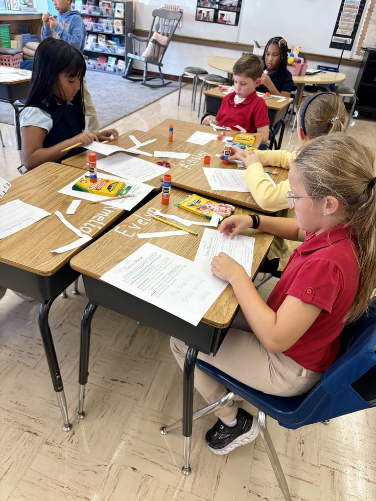 A group of elementary school students sitting at clustered wooden desks in a classroom, actively working on a reading and sequencing activity about Fulton Sheen. They are cutting paper strips with scissors and using glue sticks to paste them onto worksheets alongside Crayola markers and pencils.