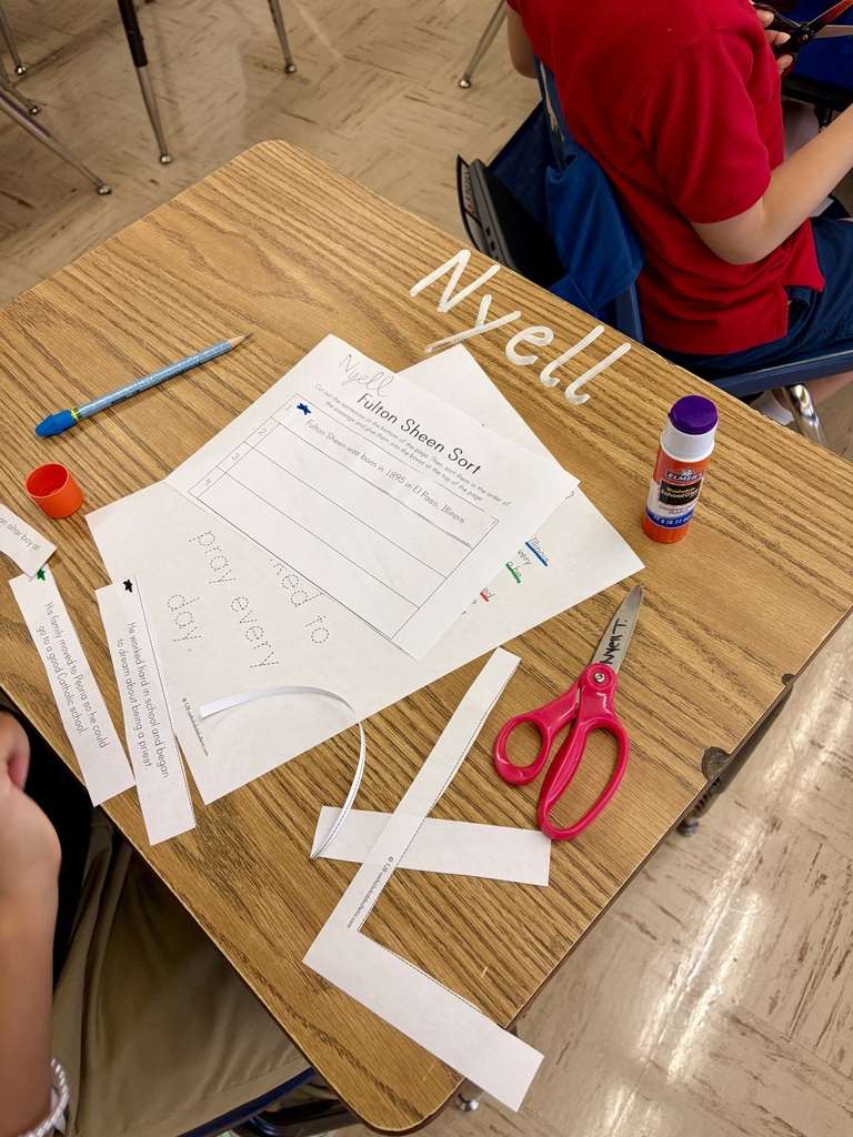 A top-down view of a student's desk set up for a classroom activity. The desk holds a worksheet titled "Fulton Sheen Sort," along with cut strips of paper containing sentences about Fulton Sheen's life, a blue glue stick, a pair of pink scissors, and a pencil.