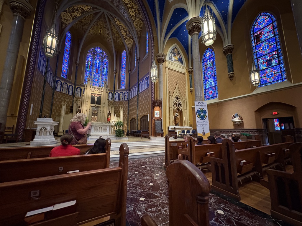 A wide-angle view from behind students seated in dark wooden pews inside a Catholic cathedral. A tour leader or teacher stands near the left foreground speaking to the group. The interior features soaring vaulted ceilings, dark red marble floors, colorful stained-glass windows along the walls, and a banner near the altar commemorating the 150th Anniversary of the Diocese of Peoria.