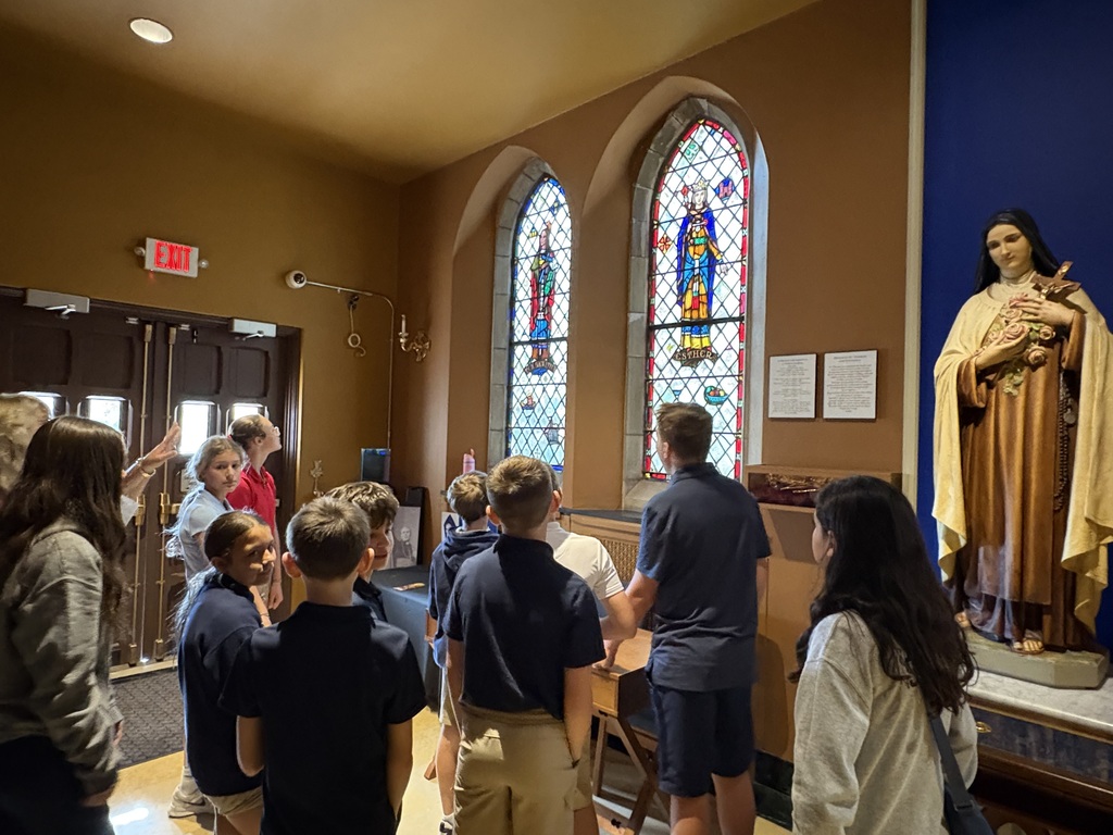 A group of students viewed from behind as they gather in a side area near the church doors. An adult chaperon or guide points toward two tall, arched stained-glass windows depicting biblical figures, including one labeled "ESTHER". To the right of the windows stands a statue of St. Thérèse holding roses and a crucifix.