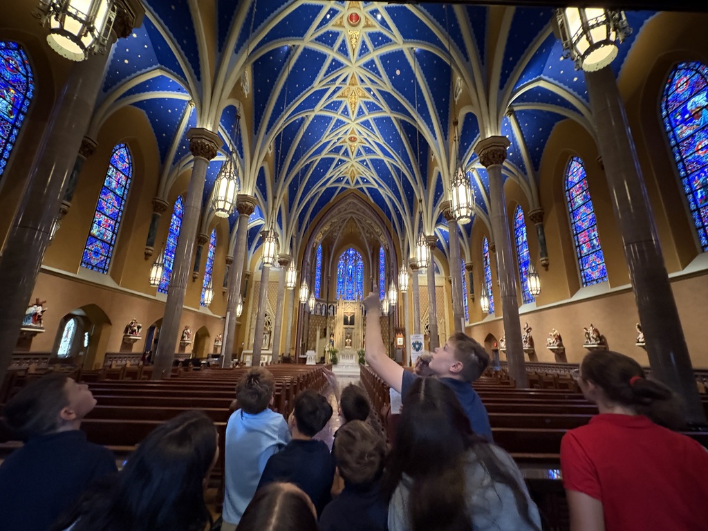 A low-angle view showing a group of students standing in the central aisle of a church, looking upward in awe. One student points toward the arched, navy blue ceiling painted with gold stars and intricate ribs. Tall stone columns, hanging lantern lights, and vibrant stained-glass windows line the sides of the sanctuary leading toward the altar in the background.