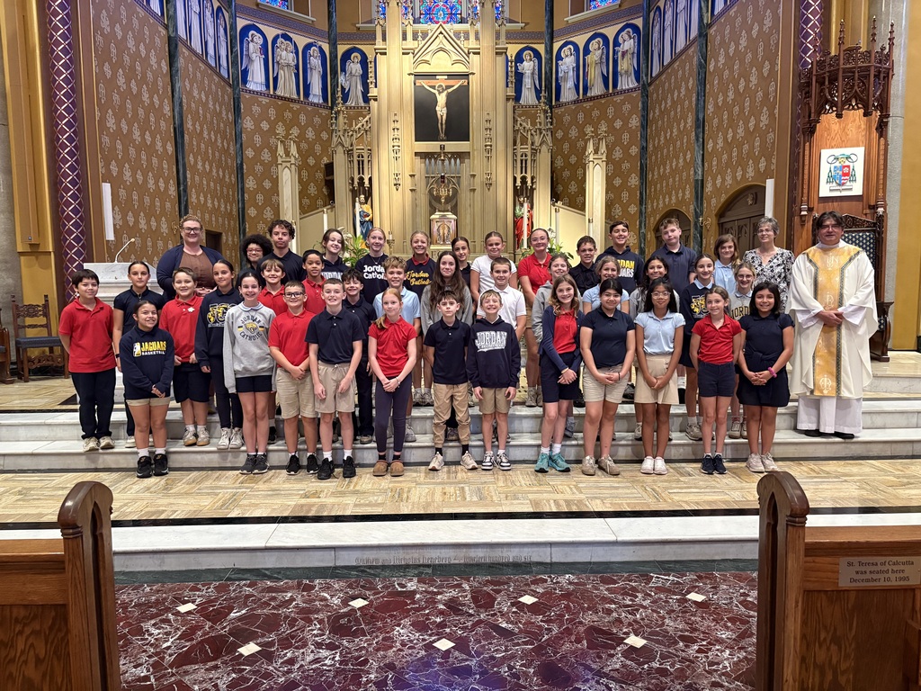 A large group of school students in red, navy blue, and grey polo shirts and sweatshirts pose for a group photo at the front altar of a Catholic church. They are standing on steps below a golden gothic-style altar adorned with statues and a crucifix. A Catholic priest in white and gold vestments stands on the far right, accompanied by adult chaperones or teachers. A plaque on the wooden pew in the foreground indicates St. Teresa of Calcutta was seated there on December 10, 1995.