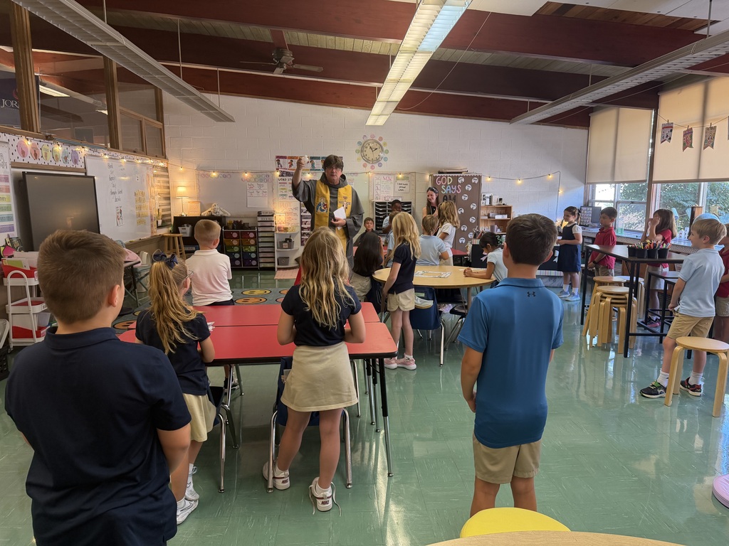 Elementary school students dressed in school uniforms (polo shirts, khakis, and skirts) stand around colorful classroom tables. At the center of the room, a Catholic priest wearing a grey habit and a priest's stole holds a small object overhead while addressing the students. The bright classroom is decorated with educational posters, a whiteboard, and string lights overhead.