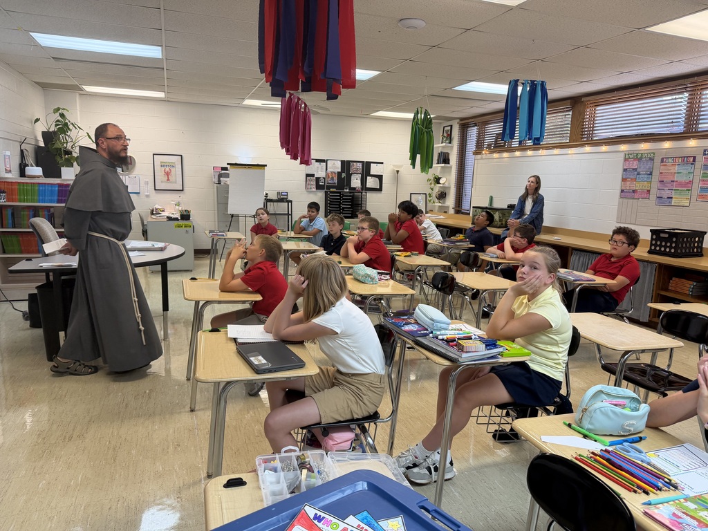 A Franciscan friar wearing a long grey habit with a cincture rope stands at the front left of a classroom speaking to students. Middle school students in red, yellow, and blue collared uniform shirts sit at individual wooden desks listening attentively, with school supplies, laptops, and notebooks open before them. Colorful paper decorations hang from the ceiling, and educational charts line the walls.