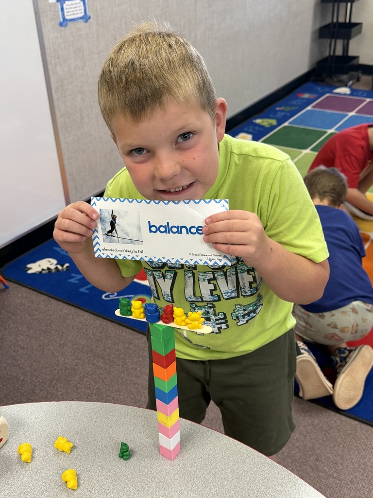 A young boy holding a word card that reads balanced above a tower of stacked blocks supporting a stick with small toy bears balanced on it.