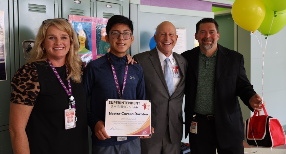 Three adults standing with a student holding an award. They are standing in front of lockers and one man is holding balloons.