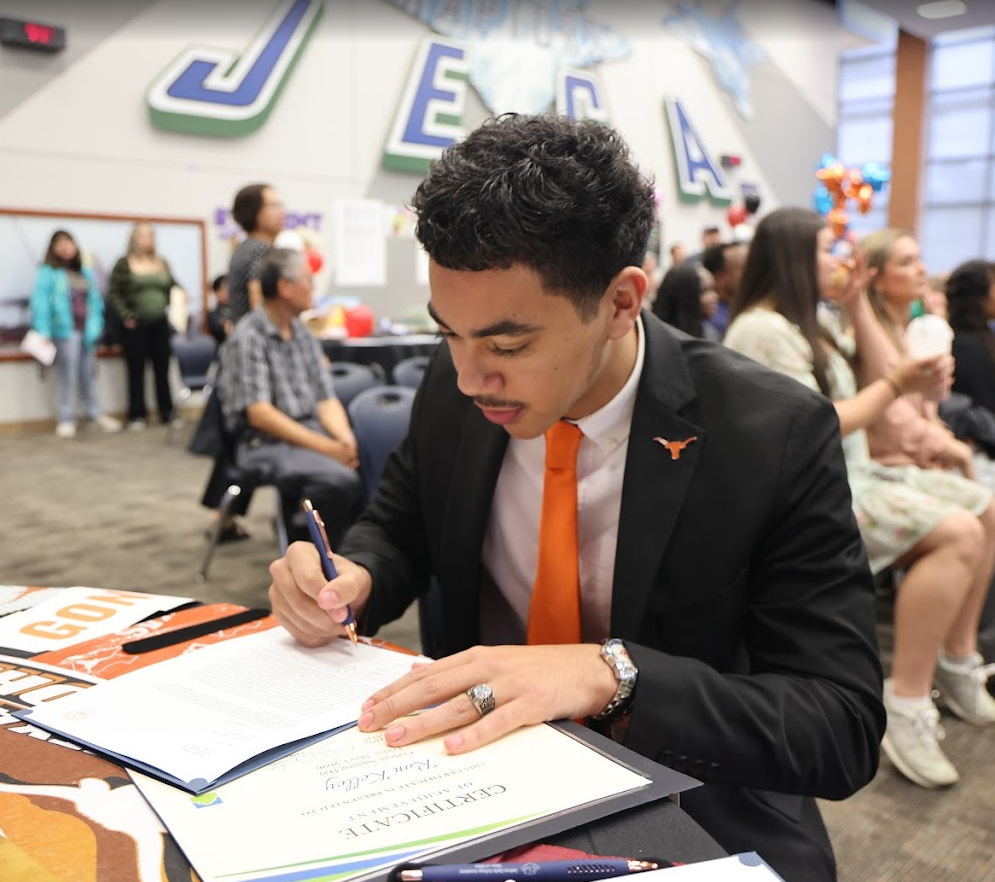 Student in a suit with a orange tie and Longhorn pin signing a paper.