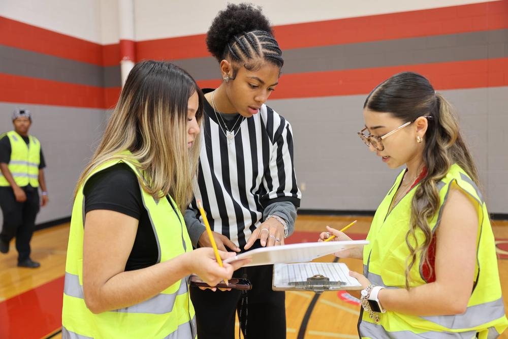 Students on a basketball court. One has on a referee uniform and the others have clipboards and yellow vests.