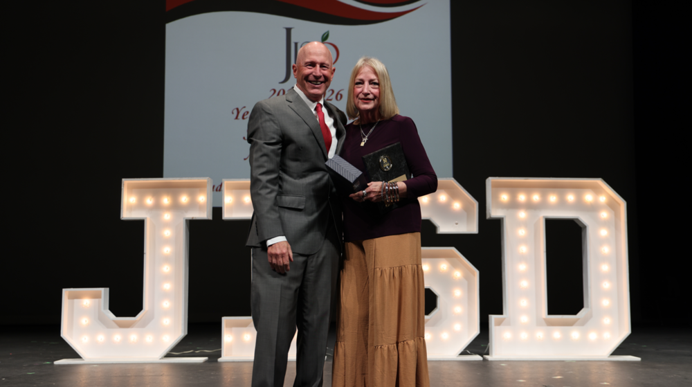 Woman with award and man in a suit in front of JISD letters