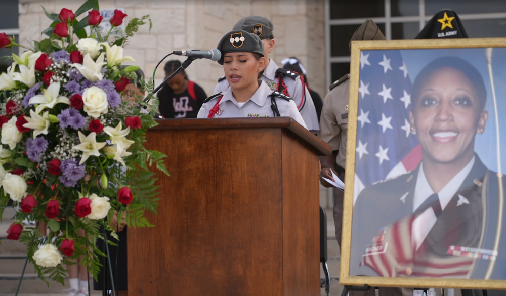 JROTC student standing behind a podium with a wreath on one side and a photo of Lt. Col. Karen Wagner on the other side.