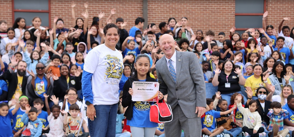 Man in suit standing with student holding an award and balloons with the principal standing beside her.Excited students stand behind them.