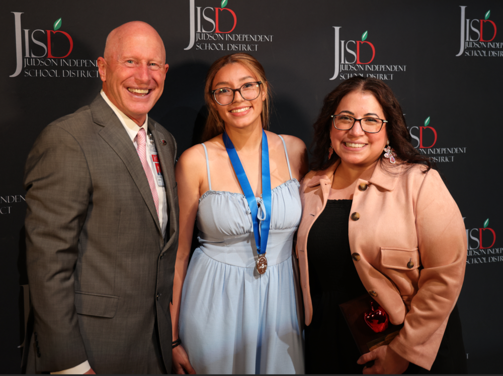 Man in suit standing with student wearing a medal and a teacher holding a plaque.