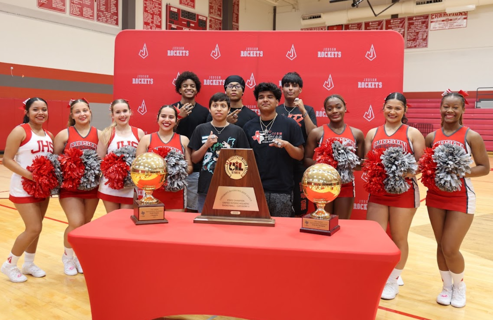 Cheerleaders standing with basketball players showing state championship rings standing in front of trophies.