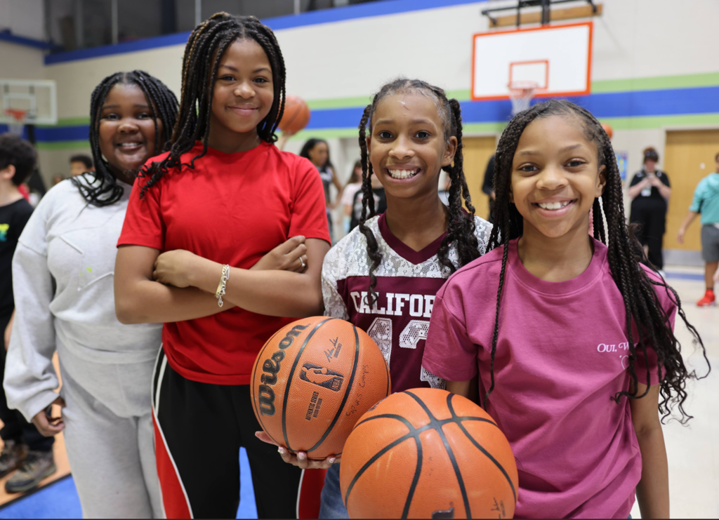 Four girls standing in a gym - two with basketballs.