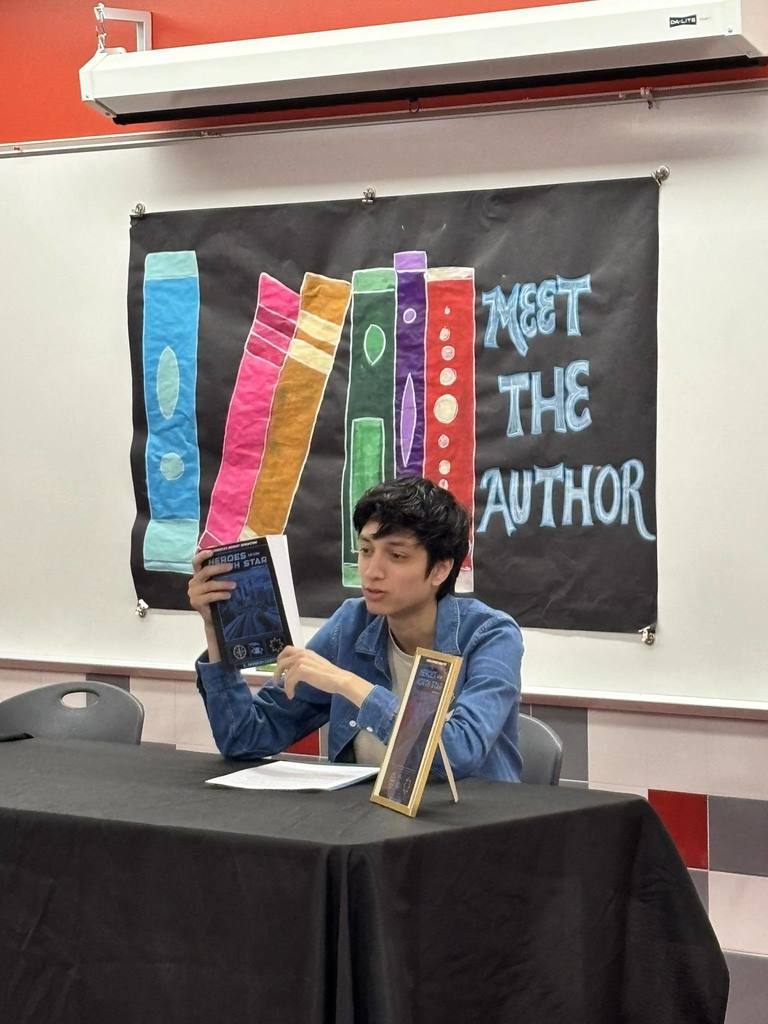 Eleazer sitting at a table holding his book while talking to an audience of students (not pictured). A "Meet the Author" banner is on the wall behind him.