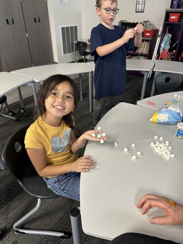 First graders using marshmallows and toothpicks to build 3D shapes in math class.