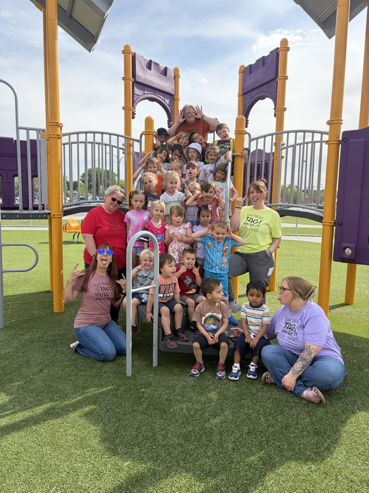 whole group photo of preschool on playground equipment outside. students and staff making silly faces