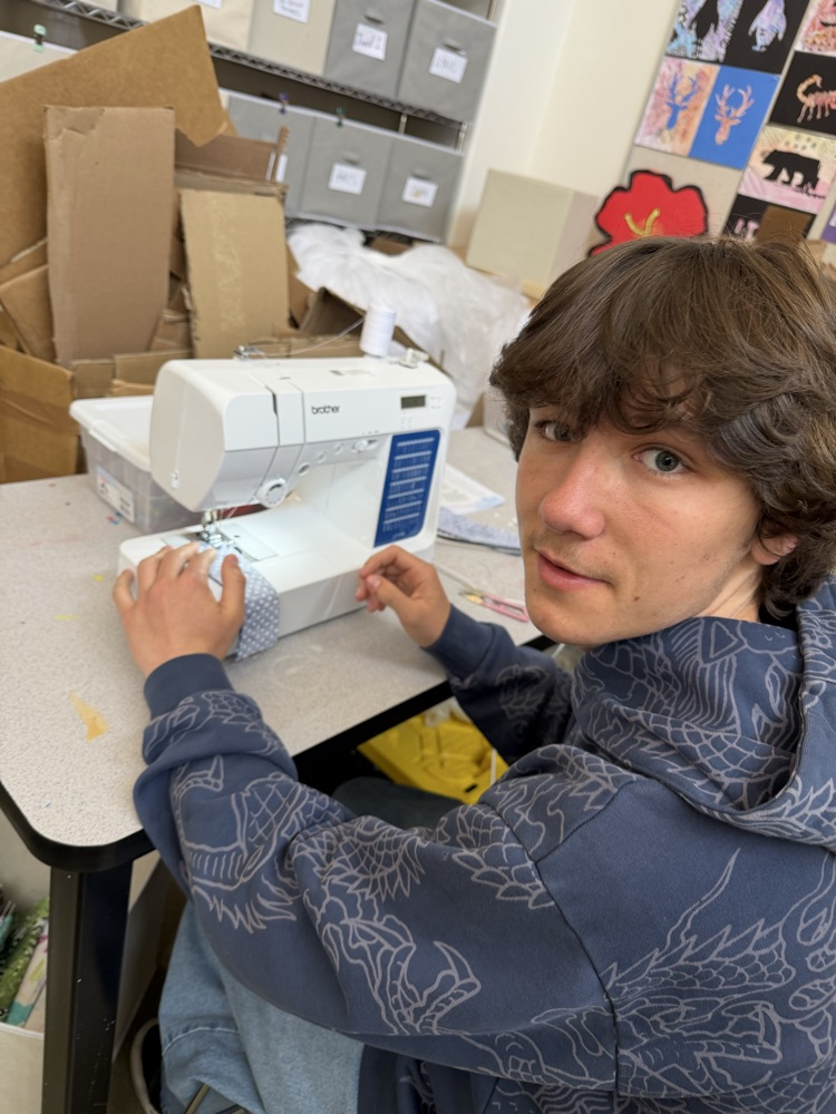 Person in a blue hoodie works on a sewing machine at a table. Background includes cardboard boxes and plastic bags.