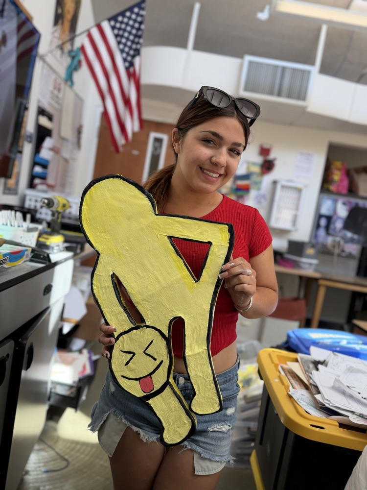 A woman holds a paper cutout of a stick figure with a happy face. She wears a red top and denim shorts. An American flag is in the background.
