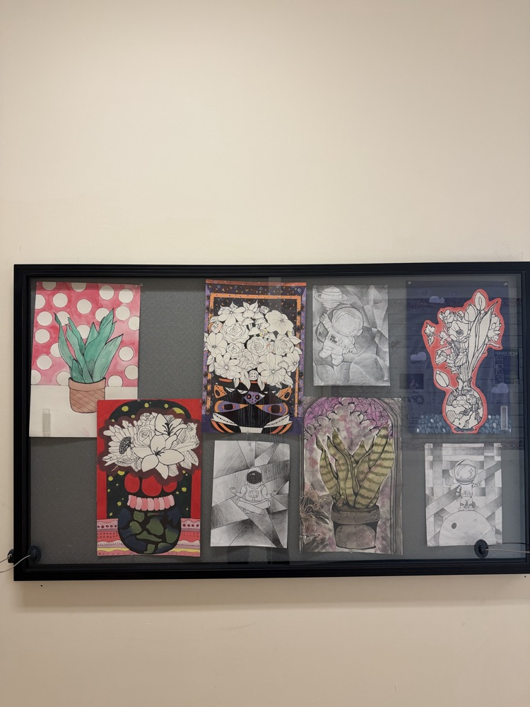Display of various art pieces, including floral and abstract designs, mounted on a wall with a black frame.