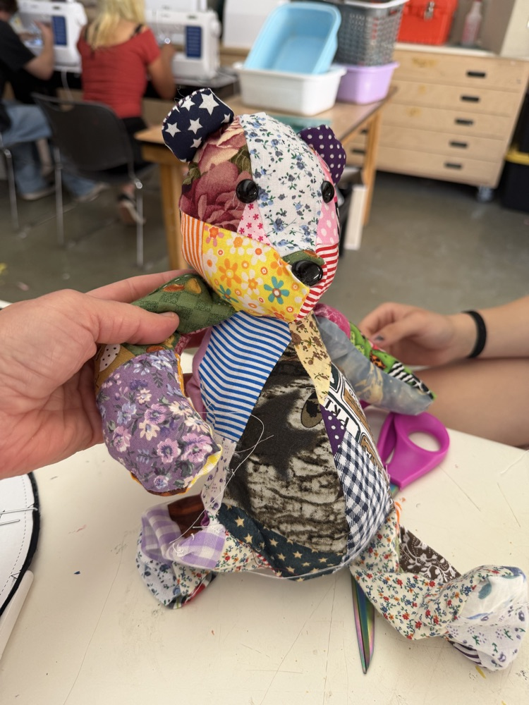 A person holds a patchwork stuffed bear with an American flag hat on a table. Other people work in the background.