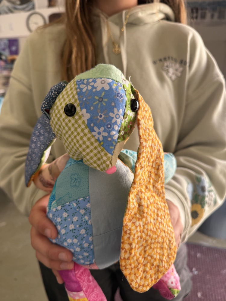 A person holds a handmade stuffed animal with a patchwork design of green, blue, pink, and yellow.