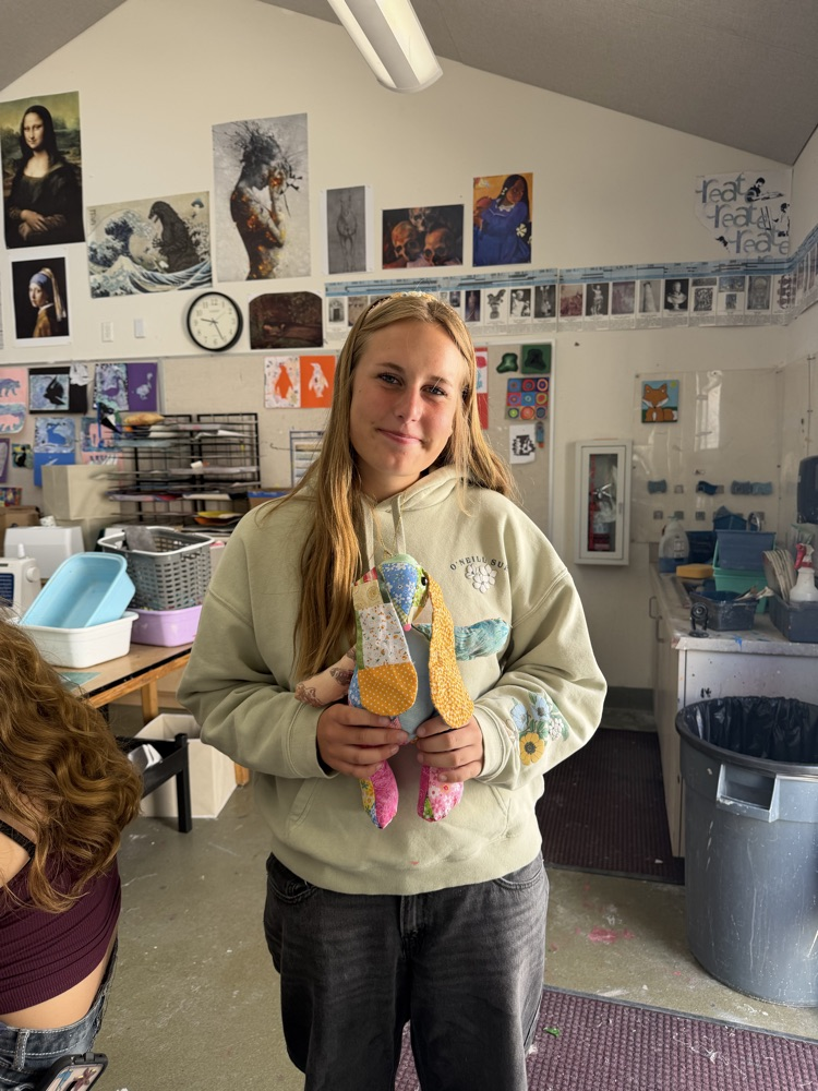 A young woman in a classroom holds a colorful doll in her hands. Behind her, various artworks adorn the walls, and a trash can is visible.