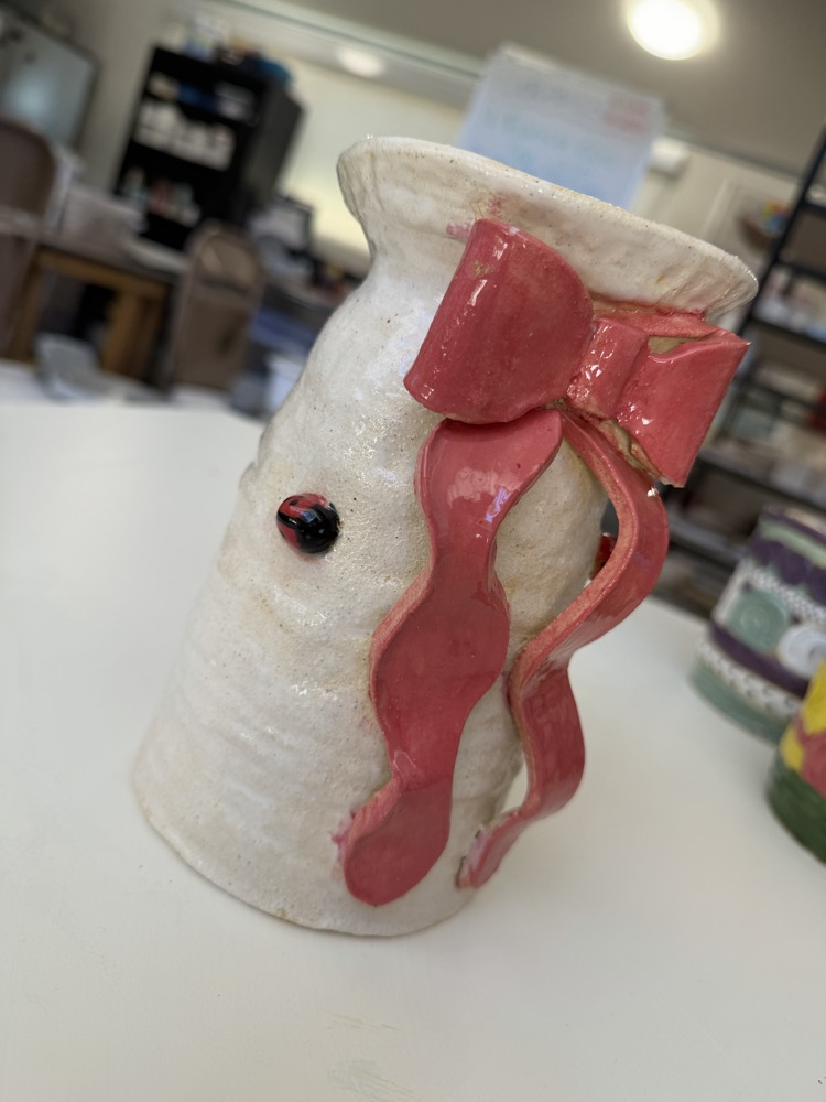 A clay vase in a workshop. It is white and has a red ribbon with a ladybug on the body.