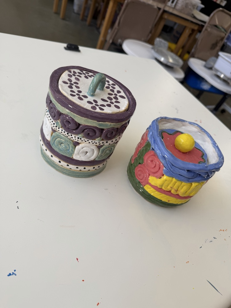 Two decorative ceramic jars on a white table. One jar is purple with floral designs, and the other is colorful with geometric shapes.