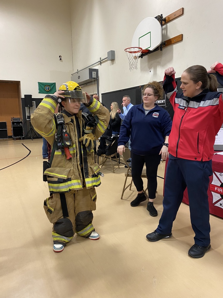 A student tries on a firefighter's uniform, and emergency personnel stand in a gymnasium.