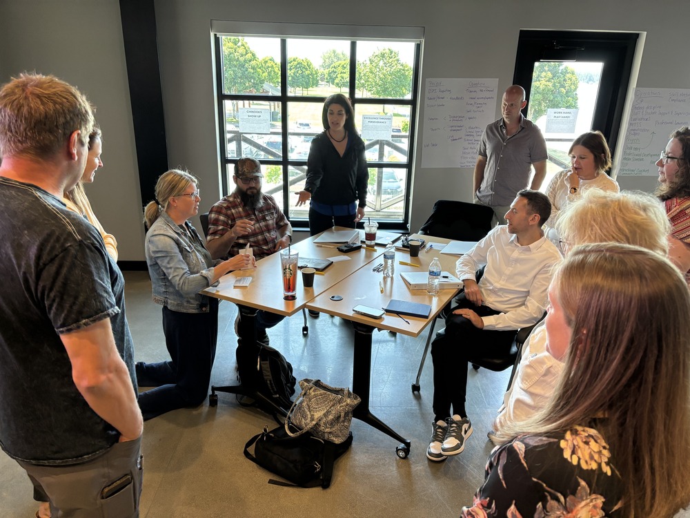 A group of people sitting around a table in a meeting room, with one person standing and gesturing while addressing the group.