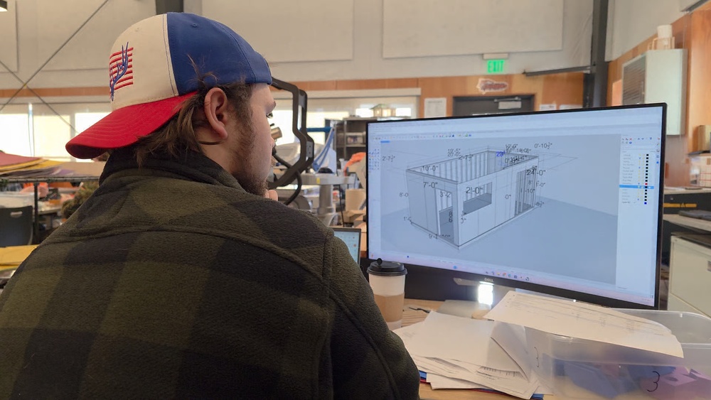 A person wearing a red baseball cap and looking at a large computer screen displaying a 3D model or blueprint.
