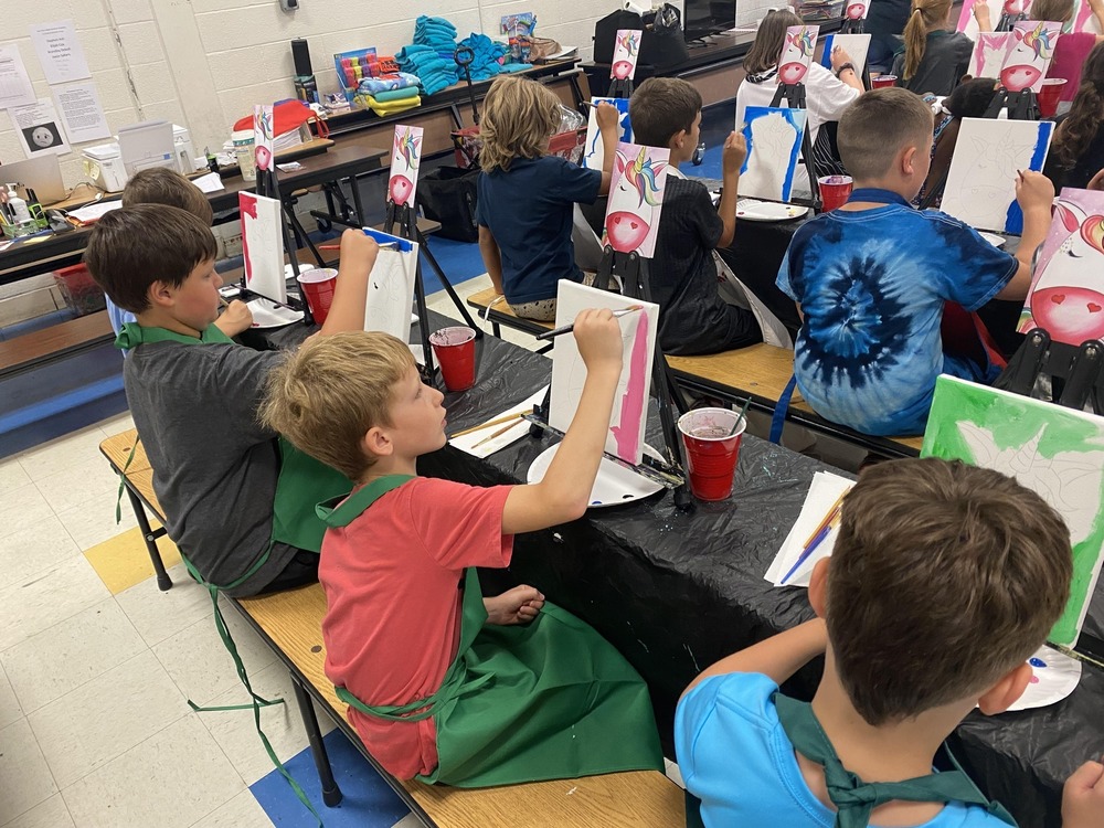 Students sitting a tables that are covered in black protective plastic. Each student is painting on a paint canvas that is set up in front of them, and they are wearing aprons.