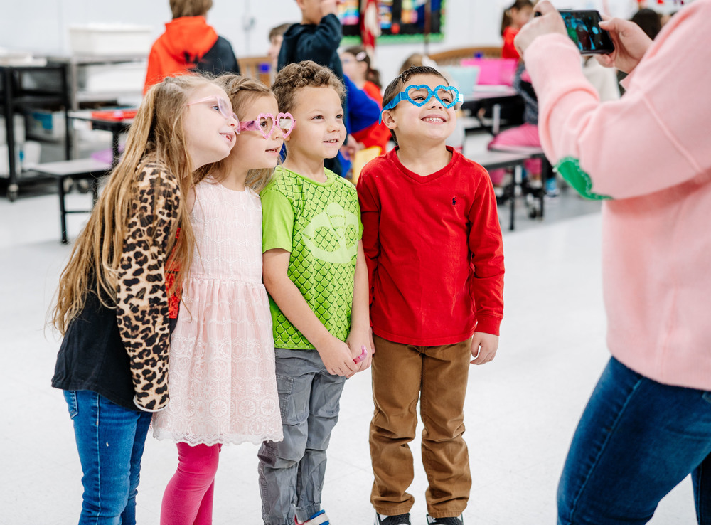 Four students are standing in the school cafeteria posing for a picture. The student's teacher is taking the picture on her cell phone. 