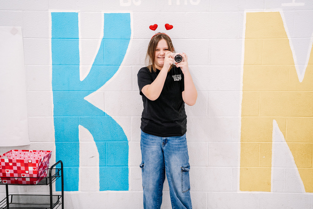 A student is holding up a digital camera and smiling. She has on a headband that has two hearts on it.