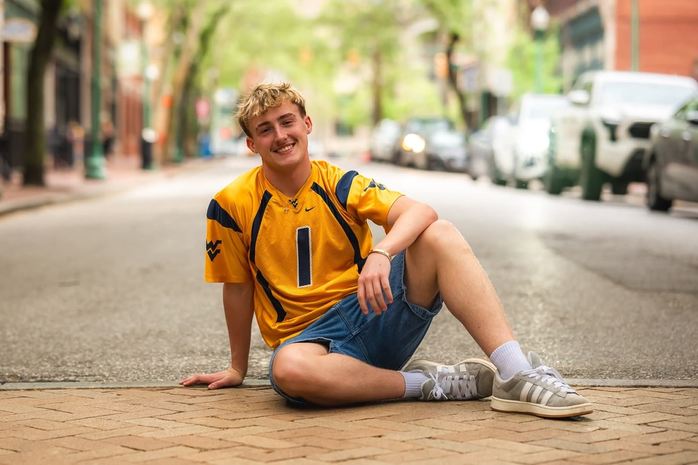 High School Senior boy with WVU jersey sitting in the road posing for a picture