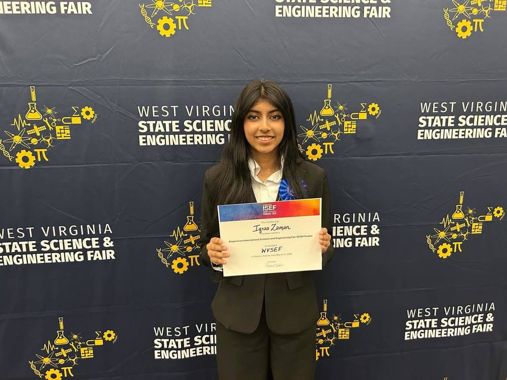 A student standing in front of the photo wall at the west virginia state science and engineering fair holding up a certificate.