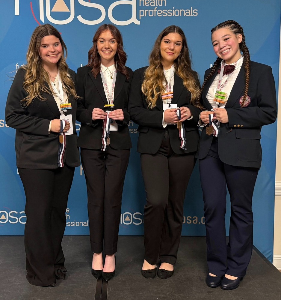 Four HOSA students are standing on stage, holding their medals they won during the competition. They are standing in front of a HOSA picture backdrop.