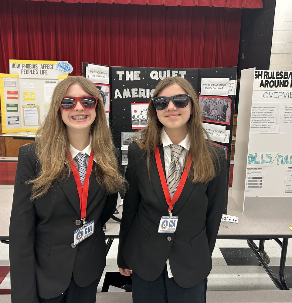 Two students dressed in black suits with a red lanyard around their necks. The badge attached to the lanyard says CIA. One student has on red sunglasses and the other has on black ones. They are standing in front of their presentation board at the social studies fair. 