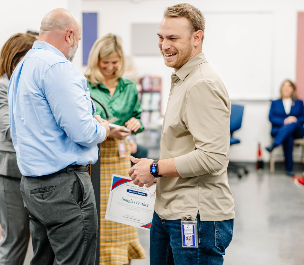 A KCS teacher is smiling after being recognized for his students success on a recent test. He is holding a certificate with his name on it.