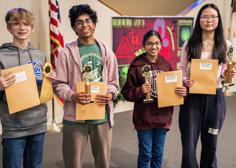 Four students standing on a stage, holding their awards from the regional math field day contest. One student, is holding a yellow ribbon and is the alternate. The other three students have trophies. 