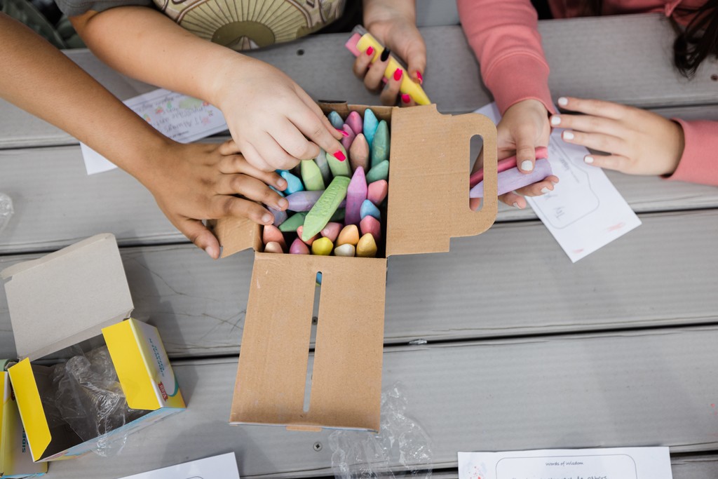 students arms reaching for chalk that is sitting in a box on top of a picnic table.
