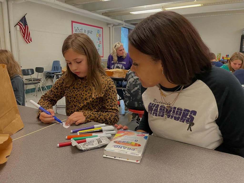 a riverside high school student looking on as an elementary student colors part of her butterfly blue.