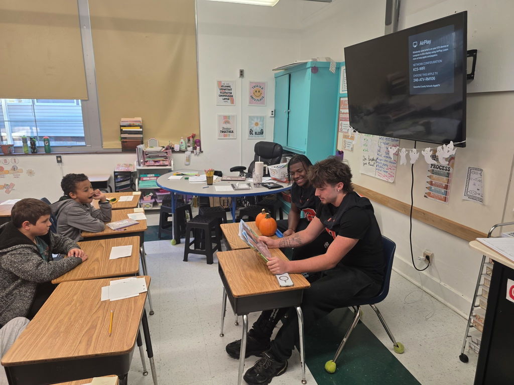 two south charleston high school football players sitting in front of an elementary classroom reading to students.