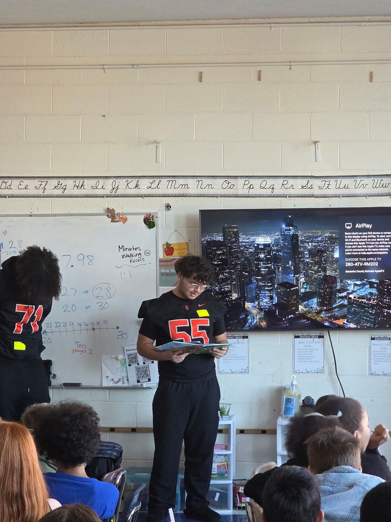 two south charleston high school football players standing in front of an elementary classroom reading to students.