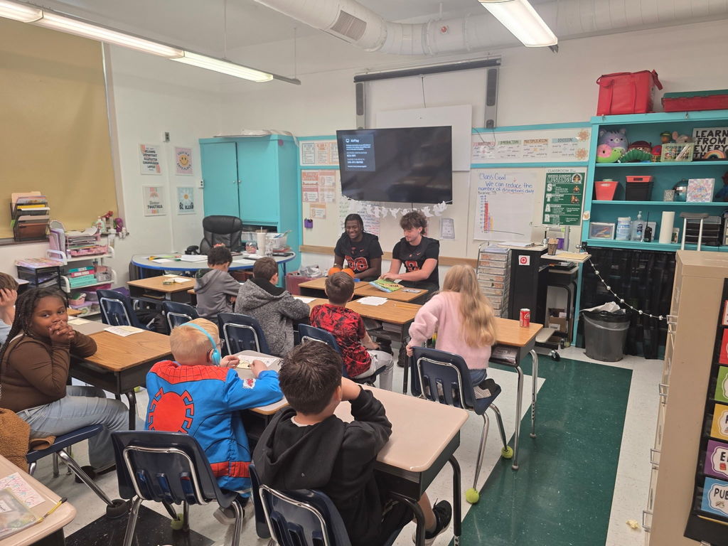 two south charleston high school football players sitting in front of an elementary classroom reading to students.