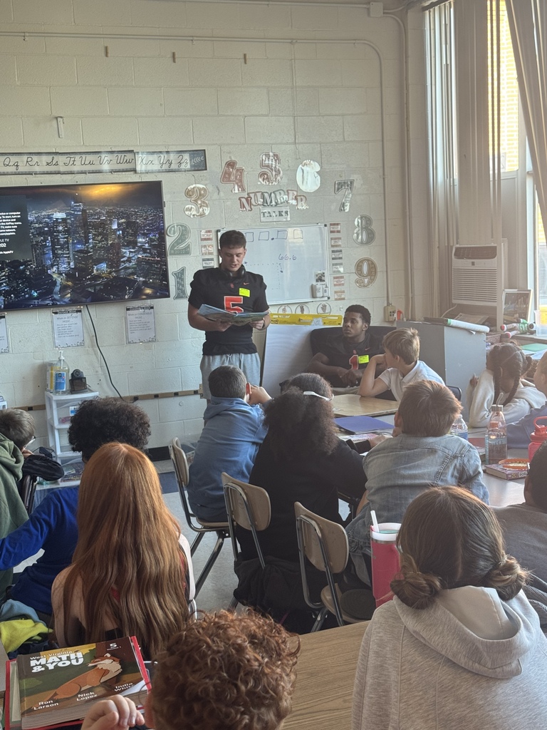 two south charleston high school football players standing in front of an elementary classroom reading to students.